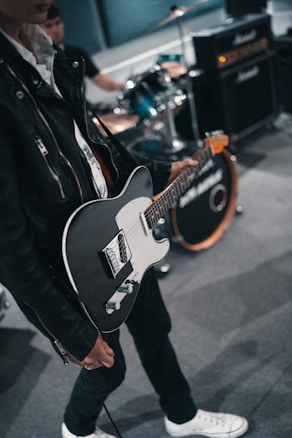 A person wearing a black leather jacket is holding a black and white electric guitar. In the background, a drummer is playing on a drum kit, and there are amplifiers visible. The setting appears to be a music studio or rehearsal space.