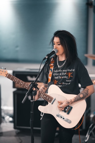 A person with tattoos is playing an electric guitar and singing into a microphone. They are wearing a dark T-shirt and a chain necklace. The background includes amplifiers and musical equipment.