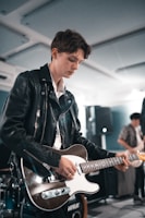 A person wearing a leather jacket is focused on playing an electric guitar. The setting appears to be indoors with another person playing a guitar in the background. The lighting is soft and ambient, suggesting a relaxed atmosphere.