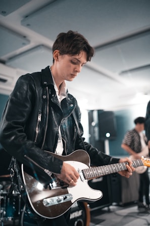 A person wearing a leather jacket is focused on playing an electric guitar. The setting appears to be indoors with another person playing a guitar in the background. The lighting is soft and ambient, suggesting a relaxed atmosphere.