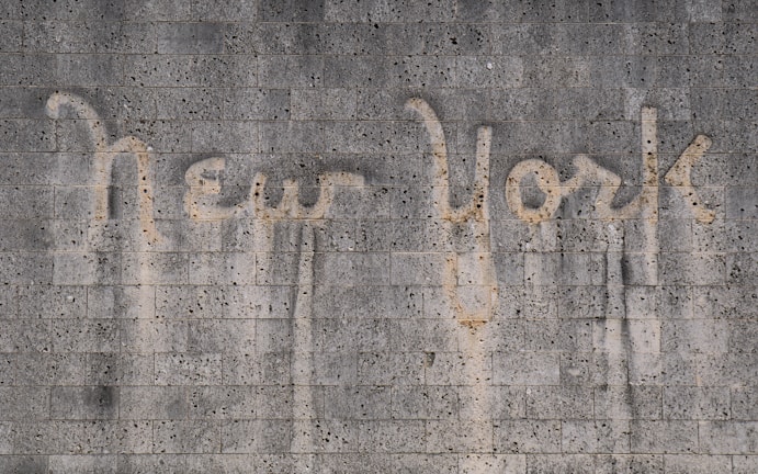 The words 'New York' are written on a textured concrete wall. The text appears to be created by a cleaning method that leaves a cleaner area in the shape of the words against the dirtier surface of the wall.