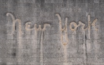 The words 'New York' are written on a textured concrete wall. The text appears to be created by a cleaning method that leaves a cleaner area in the shape of the words against the dirtier surface of the wall.
