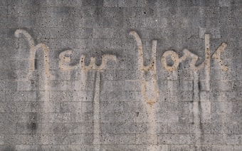 The words 'New York' are written on a textured concrete wall. The text appears to be created by a cleaning method that leaves a cleaner area in the shape of the words against the dirtier surface of the wall.