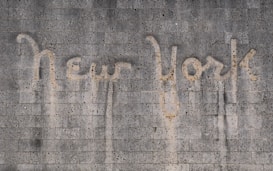 The words 'New York' are written on a textured concrete wall. The text appears to be created by a cleaning method that leaves a cleaner area in the shape of the words against the dirtier surface of the wall.
