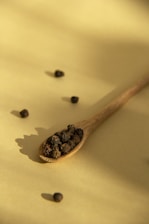 A wooden spoon filled with black peppercorns rests on a flat, light yellow surface. Scattered around the spoon are additional peppercorns, casting small, distinct shadows. The lighting creates a soft and warm ambiance with gentle shadow play.