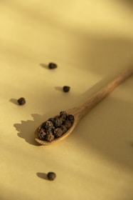 A wooden spoon filled with black peppercorns rests on a flat, light yellow surface. Scattered around the spoon are additional peppercorns, casting small, distinct shadows. The lighting creates a soft and warm ambiance with gentle shadow play.