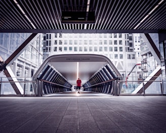A modern pedestrian bridge in an urban setting featuring sleek architectural lines. A solitary person wearing a red shirt walks across the bridge, which leads to a high-rise office building. The area is surrounded by other towering skyscrapers with reflective glass windows. The lighting is architectural, with shadows cast by the overhead structure.