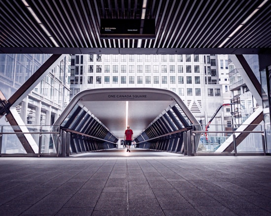 A modern pedestrian bridge in an urban setting featuring sleek architectural lines. A solitary person wearing a red shirt walks across the bridge, which leads to a high-rise office building. The area is surrounded by other towering skyscrapers with reflective glass windows. The lighting is architectural, with shadows cast by the overhead structure.