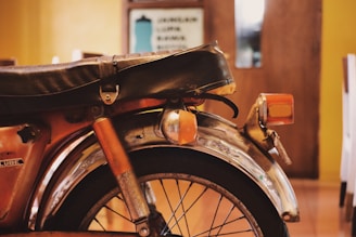 A close-up view of the rear part of a vintage motorcycle with a worn leather seat and rusty metal frame. The orange turn signal is attached near the wheel with visible wear and tear on the fender. The background features a blurred indoor setting with warm yellow and brown tones.