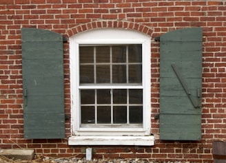 A brick wall featuring a white-framed window with an arched top and divided panes. On either side of the window are green wooden shutters that are slightly worn. The window frame appears aged, with peeling paint at the bottom. Below the window, the bricks have some weathering and scattered debris such as small rocks and twigs.