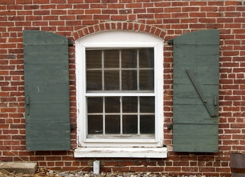 A brick wall featuring a white-framed window with an arched top and divided panes. On either side of the window are green wooden shutters that are slightly worn. The window frame appears aged, with peeling paint at the bottom. Below the window, the bricks have some weathering and scattered debris such as small rocks and twigs.