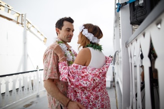 A couple dances closely on what appears to be a boat deck. They are adorned with colorful leis and the woman wears a floral headpiece and a robe with a pink floral pattern. The setting is casual and relaxed, with railings featuring decorative cut-outs visible.