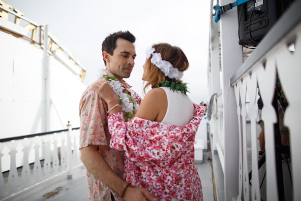 A couple dances closely on what appears to be a boat deck. They are adorned with colorful leis and the woman wears a floral headpiece and a robe with a pink floral pattern. The setting is casual and relaxed, with railings featuring decorative cut-outs visible.