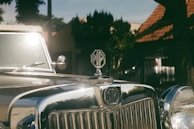 A vintage car with a shiny chrome grill and an MG emblem prominently displayed on the hood. The sunlight reflects off the car's surface, and the background features a blurred scene of houses or buildings with a red-tiled roof and greenery.