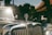 A vintage car with a shiny chrome grill and an MG emblem prominently displayed on the hood. The sunlight reflects off the car's surface, and the background features a blurred scene of houses or buildings with a red-tiled roof and greenery.