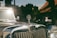 A vintage car with a shiny chrome grill and an MG emblem prominently displayed on the hood. The sunlight reflects off the car's surface, and the background features a blurred scene of houses or buildings with a red-tiled roof and greenery.