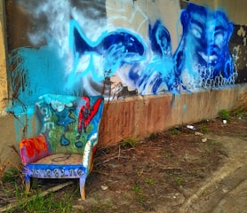 A vividly painted armchair rests against a wall adorned with vibrant blue graffiti featuring oceanic themes like a fish and abstract face. The ground is earthy with some vegetation and spray paint cans scattered nearby.