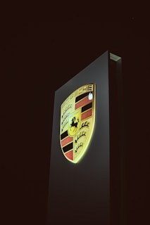 A vertical sign displays an illuminated Porsche logo against a dark background. The emblem features a combination of red, black, and gold colors with a horse in the center and the name 'Stuttgart' beneath it.