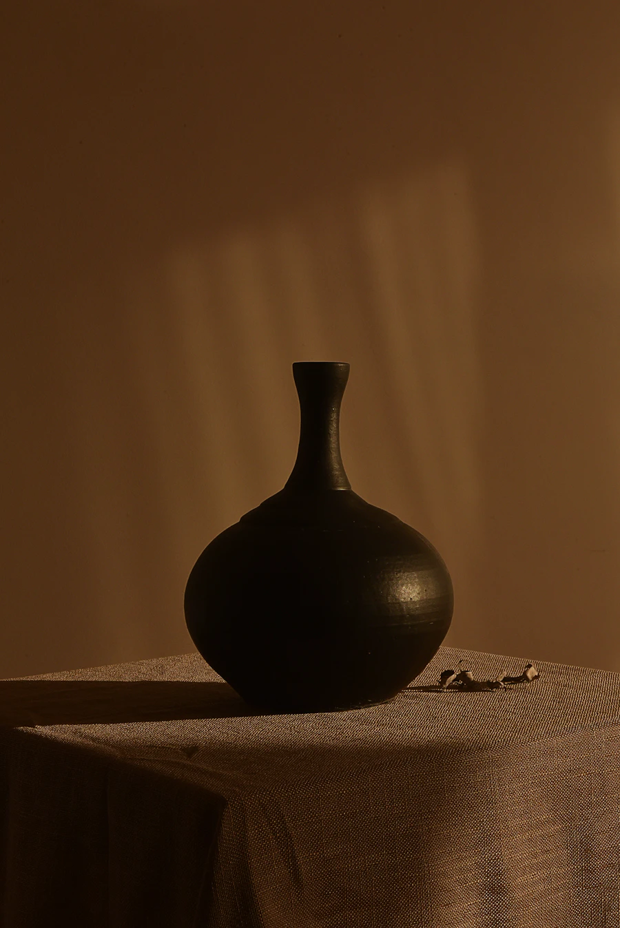 Black ceramic bud vase on a linen-draped plinth in warm amber light