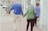 An elderly couple is walking hand in hand along a sidewalk, with the man wearing a blue sweater and beige pants, and the woman in a green sweater and black pants. They are accompanied by a younger person walking slightly ahead, wearing a blue fleece. The street has parked cars, trees, and American flags on display.