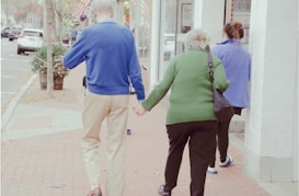 An elderly couple is walking hand in hand along a sidewalk, with the man wearing a blue sweater and beige pants, and the woman in a green sweater and black pants. They are accompanied by a younger person walking slightly ahead, wearing a blue fleece. The street has parked cars, trees, and American flags on display.