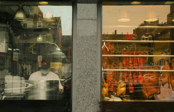 A window display of a butcher shop reveals an assortment of meats hanging on hooks, including whole chickens and cuts of red meat. The scene inside the shop shows a person wearing a face mask, possibly working, while the reflection in the glass reveals buildings and street elements outside.