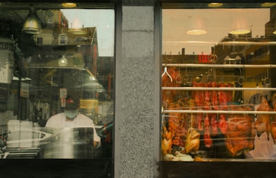 A window display of a butcher shop reveals an assortment of meats hanging on hooks, including whole chickens and cuts of red meat. The scene inside the shop shows a person wearing a face mask, possibly working, while the reflection in the glass reveals buildings and street elements outside.