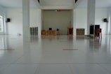 A large, empty, clean room with a tiled floor. At the far end, there is a table covered with a beige cloth, a black chair, and some items such as a fan, books, and a small podium. There are two large columns in the room and a digital clock display above the center of the back wall. Speakers are mounted on the walls. Several rolled-up mats are leaning against the walls on either side.