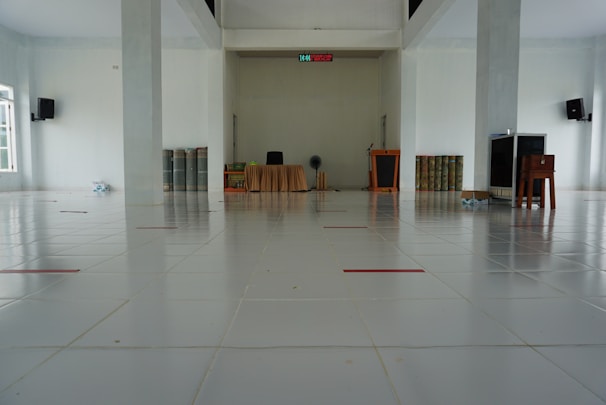 A large, empty, clean room with a tiled floor. At the far end, there is a table covered with a beige cloth, a black chair, and some items such as a fan, books, and a small podium. There are two large columns in the room and a digital clock display above the center of the back wall. Speakers are mounted on the walls. Several rolled-up mats are leaning against the walls on either side.