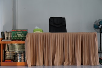 A neatly arranged desk with a beige tablecloth and a black office chair is set against a plain wall. To the left, a wooden shelf crammed with books, including a prominently placed green volume titled 'Tafsir Ibnu Katsir'. On the right, a stand fan adds a touch of comfort.
