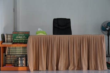 A neatly arranged desk with a beige tablecloth and a black office chair is set against a plain wall. To the left, a wooden shelf crammed with books, including a prominently placed green volume titled 'Tafsir Ibnu Katsir'. On the right, a stand fan adds a touch of comfort.