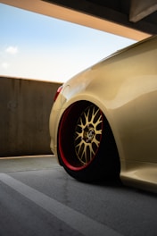 A close-up view of a sleek, customized car wheel with a gold alloy rim and red inner detailing, set against a muted urban concrete backdrop. The car's body is a smooth, shiny beige color, reflecting light subtly. The scene is under a partially covered parking area on a clear day.