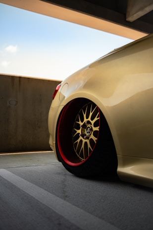 A close-up view of a sleek, customized car wheel with a gold alloy rim and red inner detailing, set against a muted urban concrete backdrop. The car's body is a smooth, shiny beige color, reflecting light subtly. The scene is under a partially covered parking area on a clear day.