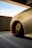 A close-up view of a sleek, customized car wheel with a gold alloy rim and red inner detailing, set against a muted urban concrete backdrop. The car's body is a smooth, shiny beige color, reflecting light subtly. The scene is under a partially covered parking area on a clear day.