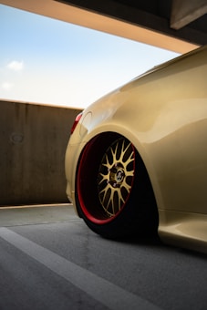 A close-up view of a sleek, customized car wheel with a gold alloy rim and red inner detailing, set against a muted urban concrete backdrop. The car's body is a smooth, shiny beige color, reflecting light subtly. The scene is under a partially covered parking area on a clear day.