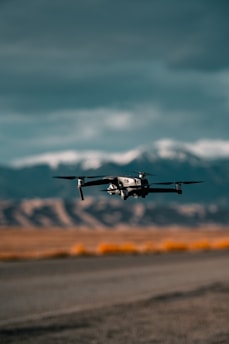 A drone is flying close to the ground with a mountainous backdrop under a cloudy sky. The landscape includes dry, rocky terrain with sparse vegetation.