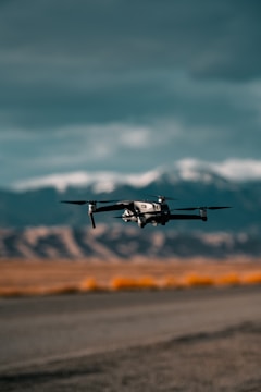 A drone is flying close to the ground with a mountainous backdrop under a cloudy sky. The landscape includes dry, rocky terrain with sparse vegetation.