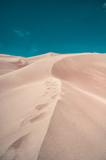 A hidden mountain trail winding through soft beige sand dunes under a clear blue sky.
