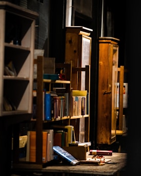 A cozy interior scene with wooden bookshelves filled with books. Warm lighting accentuates the texture of the wood, creating a rustic and inviting atmosphere. Various books and small objects are scattered on a table, adding to the eclectic charm.