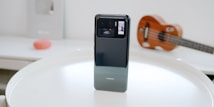 A modern smartphone is placed upright on a round white table. The phone features a glossy, reflective back with a black, square camera module at the top, which houses multiple lenses and a small display screen. In the blurred background, a wooden ukulele and a red circular object can be seen on a white surface.