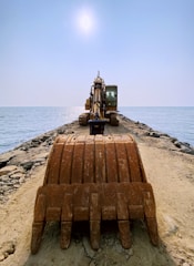 An excavator is positioned at the end of a rocky pier extending into the ocean, under a bright, clear sky. The view is from behind the large earth-moving bucket, emphasizing the machinery as it faces the sea.