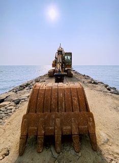 An excavator is positioned at the end of a rocky pier extending into the ocean, under a bright, clear sky. The view is from behind the large earth-moving bucket, emphasizing the machinery as it faces the sea.