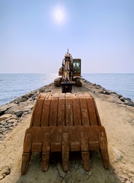 An excavator is positioned at the end of a rocky pier extending into the ocean, under a bright, clear sky. The view is from behind the large earth-moving bucket, emphasizing the machinery as it faces the sea.