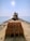 An excavator is positioned at the end of a rocky pier extending into the ocean, under a bright, clear sky. The view is from behind the large earth-moving bucket, emphasizing the machinery as it faces the sea.