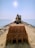 An excavator is positioned at the end of a rocky pier extending into the ocean, under a bright, clear sky. The view is from behind the large earth-moving bucket, emphasizing the machinery as it faces the sea.