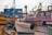 Multiple colorful fishing boats are docked at a waterfront with various names and numbers visible on their sides. Seagulls perch on some of the boats, and the dock is lined with weathered posts and ropes. The scene is calm and serene, with overcast skies in the background.