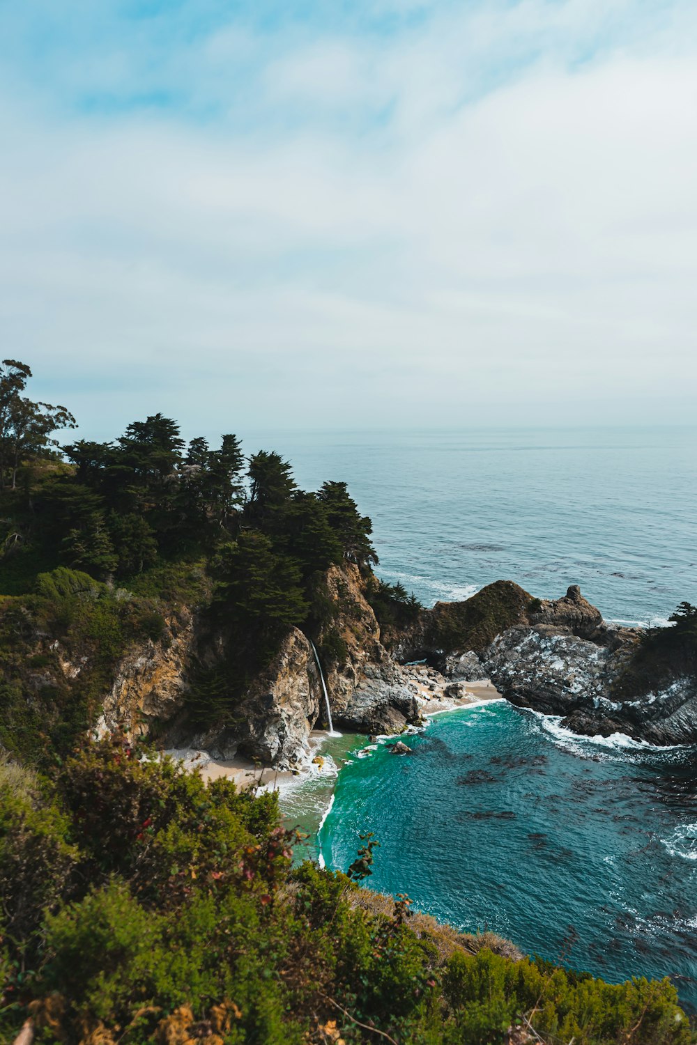 green trees on brown rocky mountain beside blue sea under white clouds during daytime