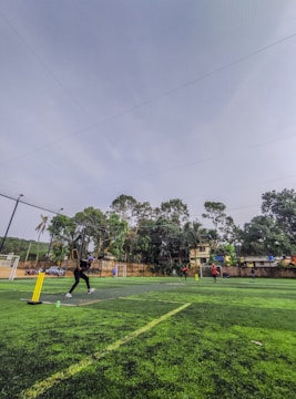 A group of people are playing a game of cricket on a grassy field surrounded by trees and a few buildings. The player on the left is batting, as another person bowls from the right. The playing area is enclosed by a net and contains cricket stumps and a pitch.