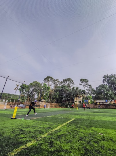 A group of people are playing a game of cricket on a grassy field surrounded by trees and a few buildings. The player on the left is batting, as another person bowls from the right. The playing area is enclosed by a net and contains cricket stumps and a pitch.
