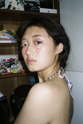 A woman with glittery makeup and tear-like decorations on her face poses in front of a shelf filled with various small objects and books. Her hair is styled in loose waves, and she wears a chain-link strap top. The background includes cluttered shelves with colorful miniature cars and visible electronics on the top shelf.